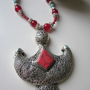 Peut inclure: Un collier en argent avec un grand pendentif orné d'une pierre rouge et de motifs complexes. Le collier est composé de perles rouges et d'argent.