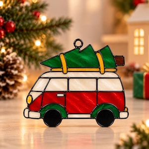 May include: A stained glass ornament shaped like a vintage van with a green Christmas tree strapped to the roof. The van features a white upper body, a red lower body, green wheel arches, black wheels, and a yellow headlight.