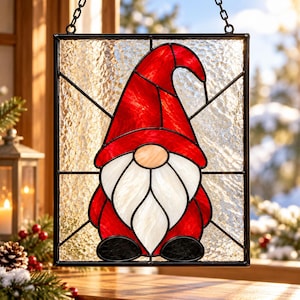 May include: A rectangular stained glass suncatcher featuring a gnome with a tall red hat, a white beard, and black shoes. The gnome is centered against a background of textured clear glass panels framed in black metal, with a hanging chain attached.