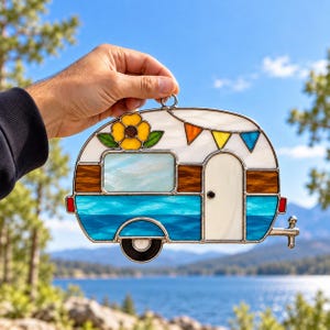 May include: A stained glass suncatcher shaped like a vintage camper trailer. The design features a blue and white body, a brown stripe, a yellow flower, and colorful triangular bunting flags. It has a metal loop for hanging.