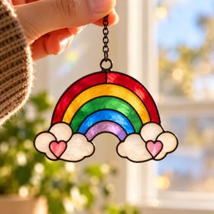 May include: A stained glass suncatcher shaped like a rainbow with red, orange, yellow, green, blue, and purple arcs. The rainbow rests on two white clouds, each featuring a small pink heart at its center. A black metal chain is attached to the top.