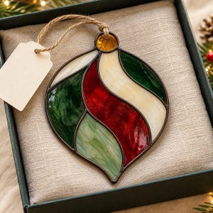 May include: A teardrop-shaped stained glass ornament featuring curved panels of deep red, forest green, light green, and cream-colored glass held together by dark metal solder lines, topped with a small amber glass accent and a jute twine hanging loop.