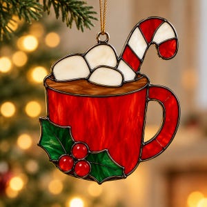 May include: A stained glass Christmas ornament shaped like a red mug filled with white marshmallows and a red and white striped candy cane. The mug features a green holly leaf and red berry accent on the front and hangs from a gold string.
