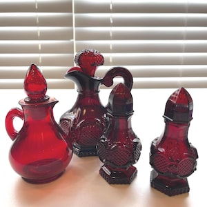 May include: A set of four ruby red glass pieces including two cruets with pointed stoppers and handles, and two shakers with hexagonal bases. Each piece features a textured, pressed glass pattern with circular and diamond-shaped motifs.