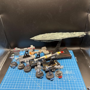 May include: A collection of miniature plastic starship models displayed on black stands. The set includes one large grey capital ship and several smaller vessels in various colours, including red, white, and orange, arranged on a blue cutting mat.