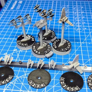 Puede incluir: Una colección de naves espaciales en miniatura de plástico gris claro sin pintar y bases de juego circulares negras con números blancos impresos. Varias naves están montadas en soportes verticales, mientras otras siguen en el marco.
