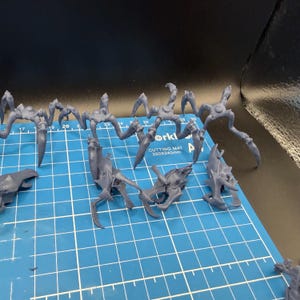 May include: A collection of unpainted grey resin miniature figures featuring multi-legged insectoid creatures and separate head components. The figures are arranged on a blue cutting mat with white grid lines and text reading 320x240mm.