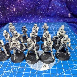 May include: A collection of fifteen miniature stormtrooper figurines, each painted in white armor with black detailing. The figures are posed in various standing and aiming positions, each mounted on a circular black base, arranged in a grid formation.