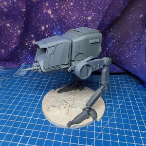 May include: A gray plastic model of a bipedal walker vehicle featuring a boxy cockpit with a front-mounted cannon, two articulated legs, and a circular base with a textured surface depicting a small figure lying on the ground.