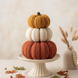 Puede incluir: Una pila de tres calabazas de ganchillo en amarillo mostaza, crema y naranja quemado, cada una con una textura acanalada y un pequeño tallo oscuro, dispuestas verticalmente sobre un soporte de pedestal de cerámica blanca.