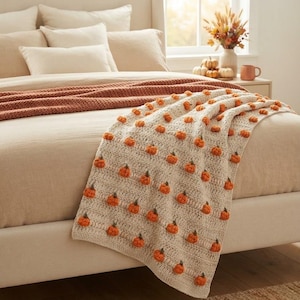 May include: A cream-colored crocheted throw blanket featuring a repeating pattern of small, raised orange pumpkins with green stems. The blanket is draped over a bed with beige bedding, showcasing the textured stitch work and pumpkin details.