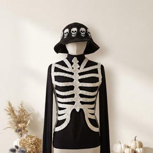May include: A black crochet bucket hat featuring a white skull pattern around the crown, worn over a black long-sleeve shirt layered with a white crochet rib cage vest that mimics the structure of a human skeleton torso.
