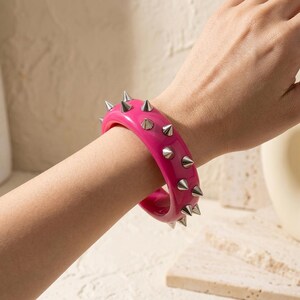 Può includere: Un bracciale rigido color rosa acceso caratterizzato da una serie di punte coniche color argento metallizzato, distanziate uniformemente e sporgenti dalla superficie esterna. Il bracciale ha una finitura liscia e lucida.