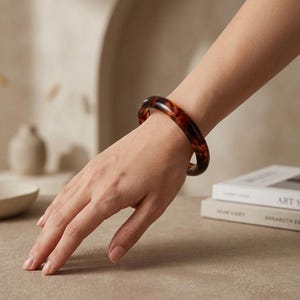 Op de afbeelding: Een gladde, ronde armband met een klassiek schildpadpatroon in wervelende tinten donkerbruin, amber en zwart. Het cirkelvormige accessoire heeft een gepolijste, glanzende afwerking en een solide, doorlopend bandontwerp.