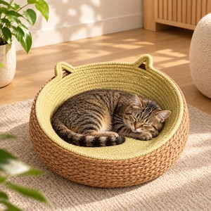 May include: A round, woven pet bed featuring a natural tan exterior and a soft, light green interior lining. The bed has two small, pointed ears extending from the top rim, and a brown tabby cat is curled up sleeping inside the circular structure.