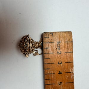 Könnte beinhalten: Ein goldfarbener Metallanhänger in Form einer kleinen, verzierten Urne oder eines Gefäßes mit filigranem Käfigdesign. Der Anhänger liegt neben einem Holzlineal, das zeigt, dass das Objekt eine Gesamtbreite von etwa 1,5 Zentimetern hat.