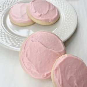 May include: Several round sugar cookies with pale pink frosting are arranged on a white plate and surface. The cookies have a light yellow base and are topped with a thick layer of pink frosting. The plate has a decorative edge.