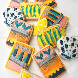 May include: A collection of decorated cookies, including sardine can shapes and lemon slices. The sardine cookies feature colorful designs with the word "Sardines" and illustrations of fish. Other cookies have blue and white patterns.