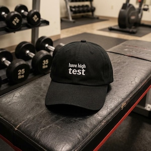 Puede incluir: Una gorra de béisbol negra con una corona estructurada y una visera curva. La parte frontal de la gorra presenta un texto bordado en blanco que dice "have high test" en una fuente minúscula y limpia. La gorra está hecha de tela negra.