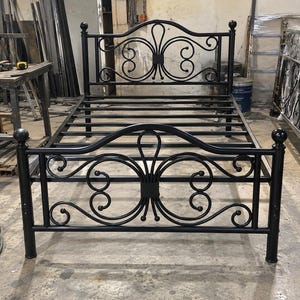 May include: A black metal bed frame featuring a headboard and footboard with symmetrical scrollwork designs. The frame includes a series of horizontal slats for mattress support and four vertical posts topped with spherical finials.