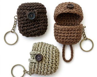 Op de afbeelding: Drie gehaakte hoesjes voor draadloze oordopjes, elk voorzien van een knoopsluiting en een metalen sleutelhangerring. Eén hoesje is donkerbruin, één is middenbruin en één is beige. Het middenbruine hoesje is open en toont de binnenkant.