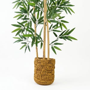Può includere: Una pianta di bambù artificiale con molteplici steli marrone chiaro e foglie verdi, contenuta in un cesto cilindrico intrecciato color giallo senape con un motivo a intreccio spesso e strutturato che copre l'intero esterno del contenitore.