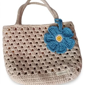 May include: A beige crochet tote bag featuring a square-patterned openwork design, two top handles, and a large blue flower charm with a yellow center attached to one handle. A small rectangular tag labeled HANDMADE is sewn on the bottom right.