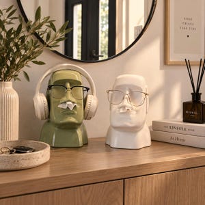 Puede incluir: Dos dispensadores de pañuelos con forma de cabeza Moai, uno en verde salvia y otro en blanco, con una nariz que dispensa los pañuelos. El dispensador verde lleva auriculares blancos y gafas de montura negra, el blanco lleva gafas de montura dorada.