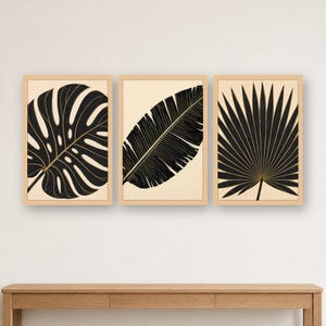Può includere: Un set di tre stampe da parete incorniciate con illustrazioni botaniche di foglie nere su sfondo beige chiaro. Ogni stampa è racchiusa in una cornice di legno chiaro e mostra rispettivamente una foglia di monstera, una di banano e una di palma.