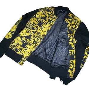 May include: A black bomber jacket featuring gold ornate scrollwork patterns on the front panels and sleeves. The jacket has a black ribbed collar, cuffs, and hem, with a black and white polka dot interior lining and Versace Jeans Couture branding.
