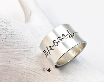 May include: A silver ring with a wide band featuring a delicate, engraved design of a vine with leaves. The ring is on a white wooden surface.