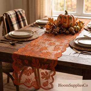 May include: A burnt orange lace table runner featuring a repeating pattern of detailed autumn leaves and a scalloped edge. The runner is draped across a wooden dining table, which is set with woven placemats, ceramic plates, and gold cutlery.