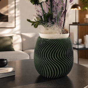 Può includere: Un vaso portapiante verde scuro con una texture a coste verticali ondulate. Il vaso ha una forma arrotondata e bulbosa e contiene un vaso interno bianco con una composizione floreale di garofani bianchi e steli di lavanda essiccata.