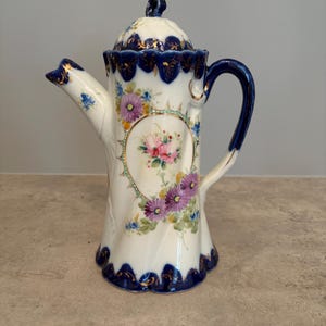 Peut inclure: Une cafetière en porcelaine au corps blanc avec une texture cannelée en spirale, ornée de bouquets de fleurs peints à la main en rose et violet. Le bord, la base, l'anse et le bec sont rehaussés d'un vernis bleu cobalt et de dorures.