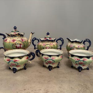 May include: A porcelain tea set featuring a teapot, a lidded sugar bowl, a creamer, and three teacups. Each piece is decorated with a light green and cream checkered pattern, pink floral motifs, and dark blue scalloped edges with gold trim.