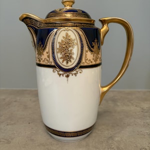 May include: A porcelain coffee pot featuring a white base, a deep blue upper section, and gold accents. The design includes a Greek key pattern, floral motifs in a central oval medallion, and a gold-toned handle, spout, and lid finial.
