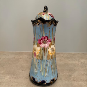 May include: A light blue ceramic jug with a lid, featuring hand-painted pink and yellow flowers, green stems, and gold leaf accents. The rim and base are decorated with a dark navy blue border and intricate gold filigree detailing.