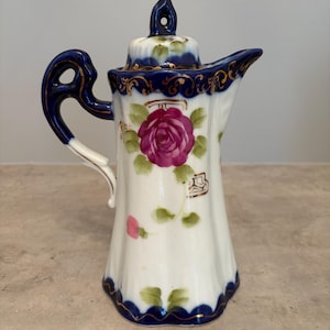 Peut inclure: Un pot à lait en porcelaine blanche au corps cannelé, doté d'un bord et d'une anse festonnés bleu foncé. La pièce est ornée d'une rose magenta peinte à la main avec des feuilles vertes, rehaussée de détails en filigrane doré sur le bord supérieur.