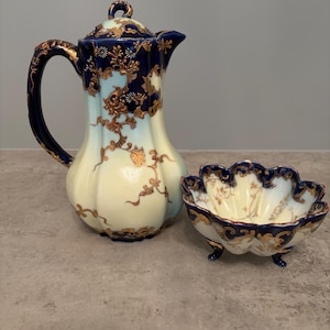May include: A porcelain coffee pot and a matching scalloped bowl featuring a cream-colored base with deep navy blue accents and intricate gold floral filigree patterns. The coffee pot has a curved handle and a lid, while the bowl has three feet.