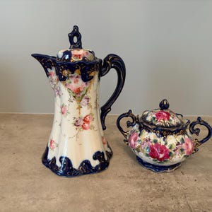 May include: A porcelain coffee pot and a matching sugar bowl, both featuring a cream-colored base with vibrant pink and purple rose floral patterns, deep navy blue accents along the rims and handles, and delicate gold trim detailing throughout.
