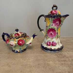 May include: A porcelain teapot and a tall coffee pot featuring a vibrant floral pattern of pink and yellow roses. Both pieces are decorated with deep blue and gold filigree trim along the edges, handles, and lids, showcasing a glossy finish.