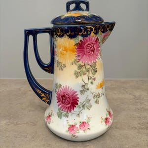 May include: A porcelain jug with a lid, featuring a cream-coloured body decorated with painted pink and yellow chrysanthemum flowers and green leaves. The handle, rim, and lid are finished in deep cobalt blue with gold-painted accents.