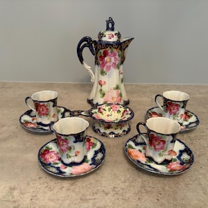 May include: A porcelain tea set featuring a tall teapot, four cups, four saucers, and a tea strainer. Each piece is decorated with vibrant pink floral patterns against a white background, accented by deep blue borders with gold-toned detailing.