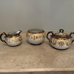 May include: A three-piece porcelain tea set featuring a creamer, an open bowl, and a lidded sugar bowl. Each piece is white with intricate gold filigree patterns, deep navy blue accents, and ornate handles on the creamer and lidded sugar bowl.