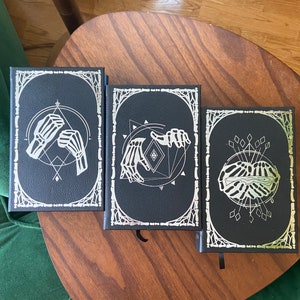May include: Three black hardcover journals with silver foil designs. The designs feature skeletal hands and geometric shapes.