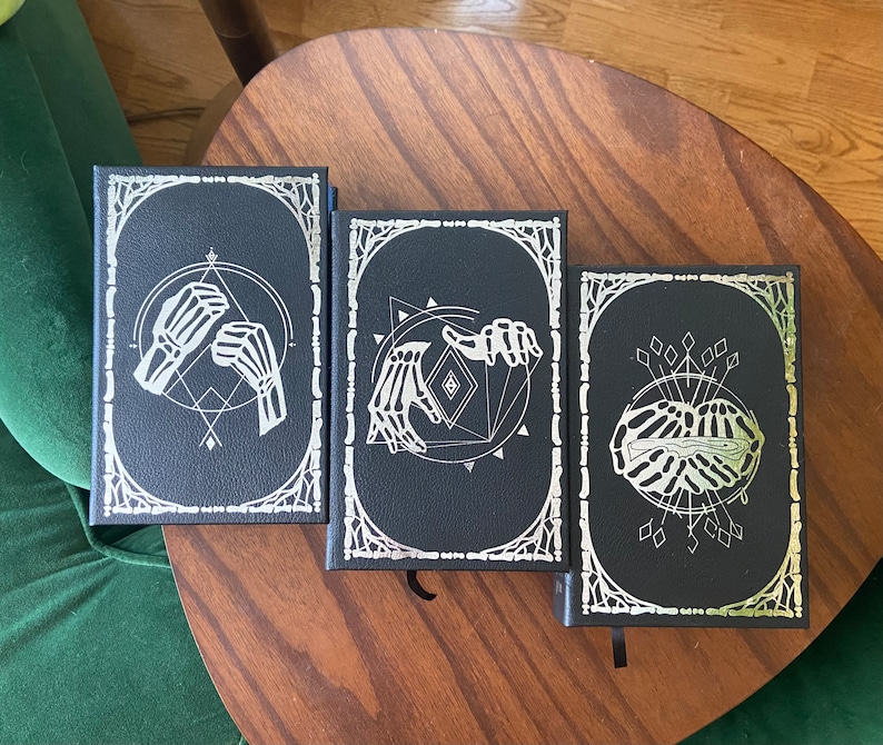 May include: Three black hardcover journals with silver foil designs. The designs feature skeletal hands and geometric shapes.