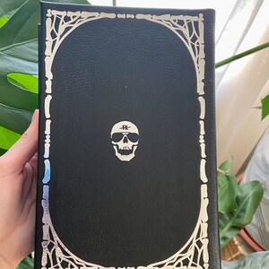 May include: Black leather-bound journal with a silver skull design and a silver border. The skull has sunglasses and a band across its forehead that says "FK".