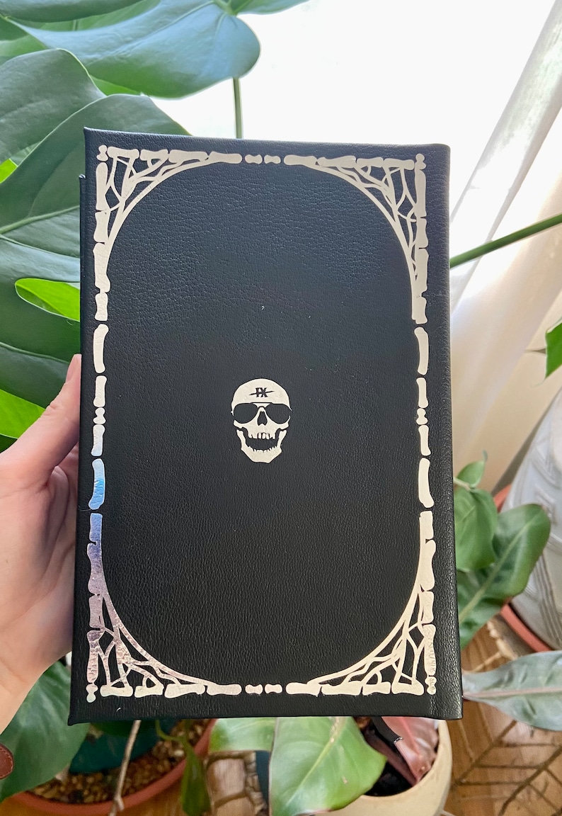 May include: Black leather-bound journal with a silver skull design and a silver border. The skull has sunglasses and a band across its forehead that says "FK".
