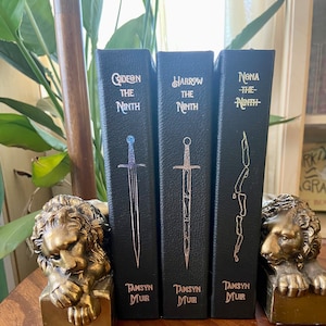 May include: Three black hardcover books with gold lettering. The titles are "Gideon the Ninth", "Harrow the Ninth", and "Nona the Ninth". The books are on a wooden surface with gold lion bookends.