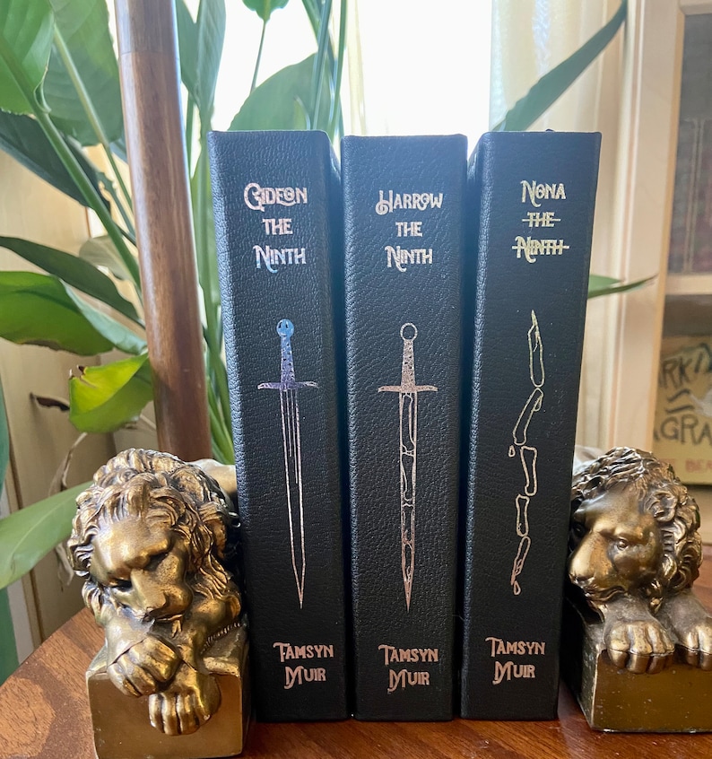 May include: Three black hardcover books with gold lettering. The titles are "Gideon the Ninth", "Harrow the Ninth", and "Nona the Ninth". The books are on a wooden surface with gold lion bookends.